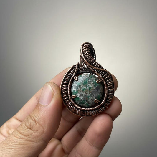 Hand holding a copper wire-wrapped pendant with a green stone against a gradient background