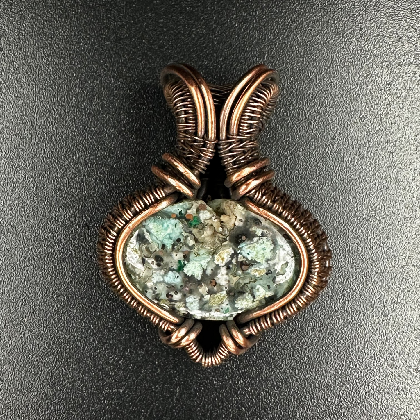 A handmade pendant featuring a natural green and white marbled stone wrapped in textured copper wire, set against a dark gray background.