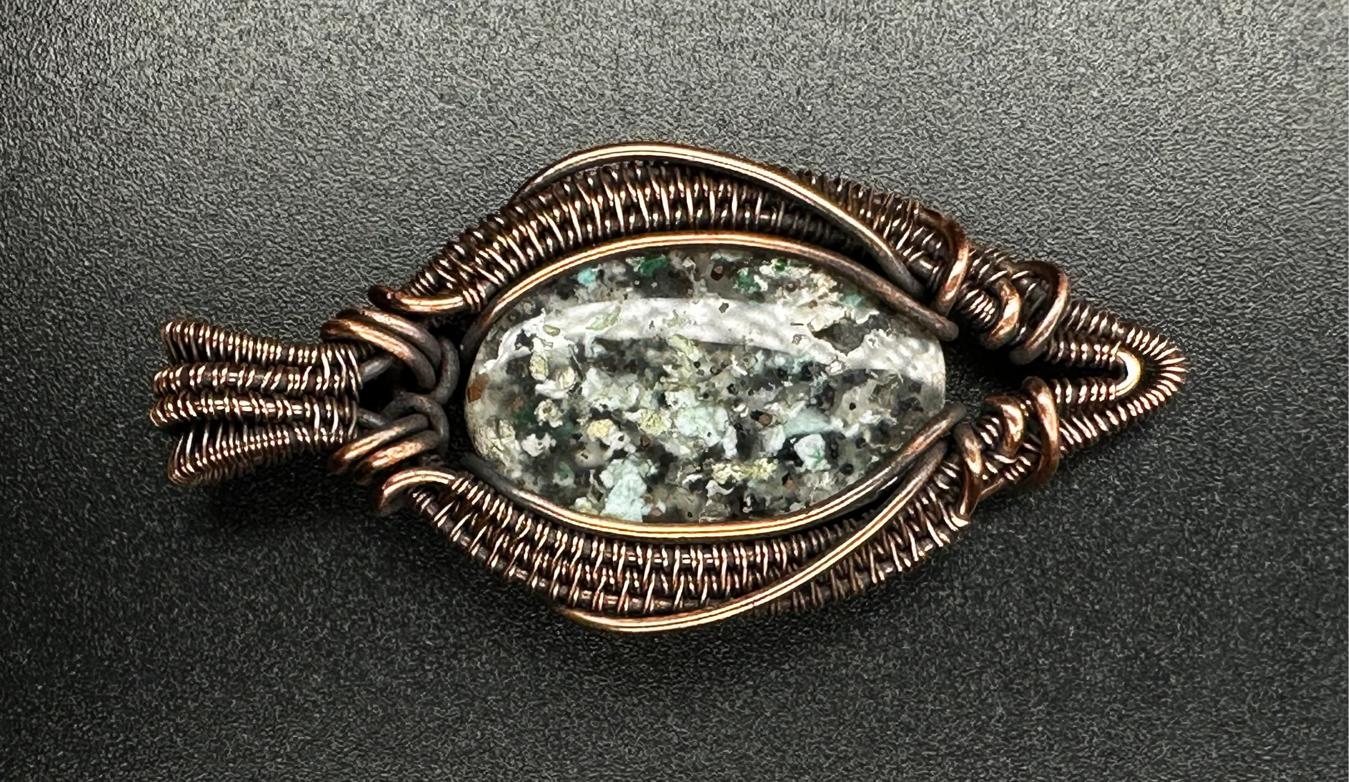 A handmade oval pendant featuring a natural marbled stone with green, white, and gray tones, wrapped in textured copper wire, set against a dark gray background.